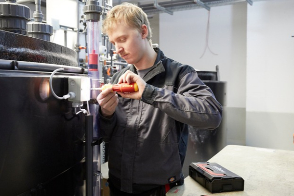 Junger Handwerker arbeitet mit einem Schraubendreher an einer technischen Anlage mit Rohren und Behältern in einem Betriebsraum.